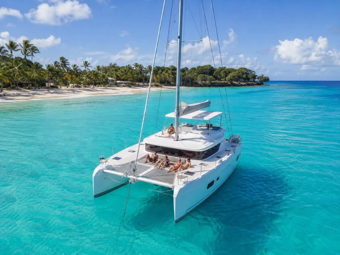 A white sailing catamaran with guests on the trampoline nets, anchored in turquoise water off a palm-lined Barbados beach
